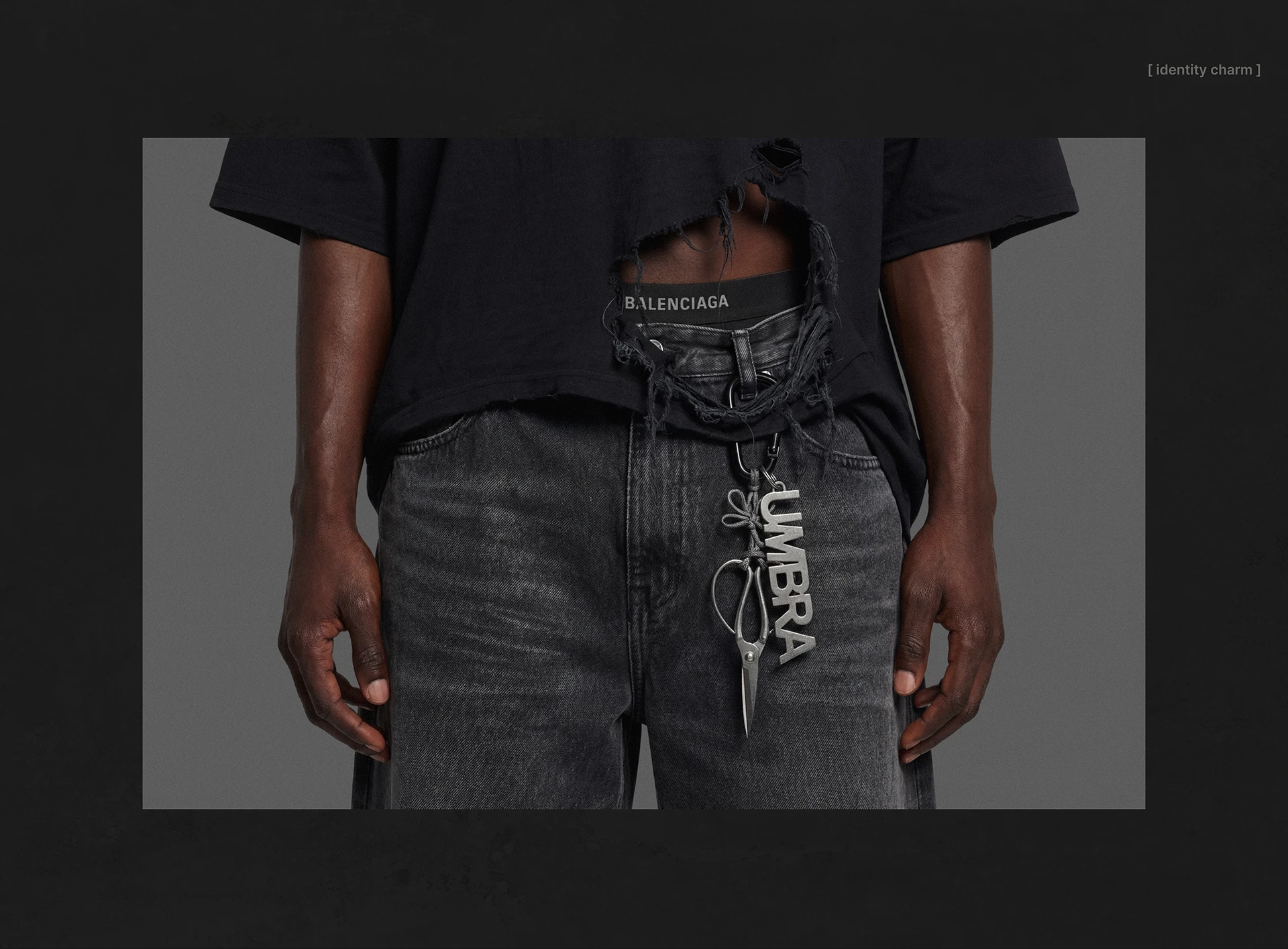 UMBRA identity charm shown as a silver branded accessory clipped to black denim, featuring the logo mark and floral-inspired metal detail in a dark editorial composition.