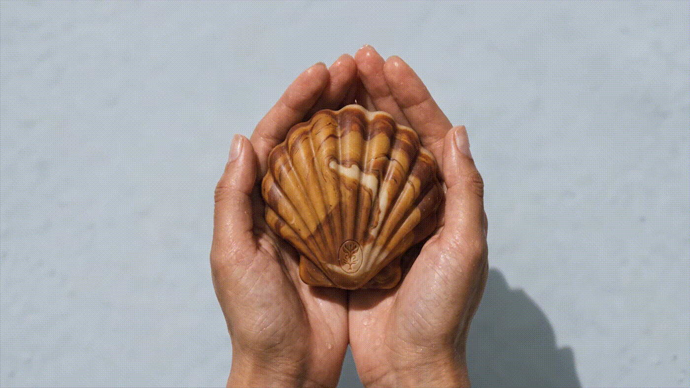 Hands holding a marbled shell-shaped NULA soap bar, highlighting the sculptural product form and natural organic texture.