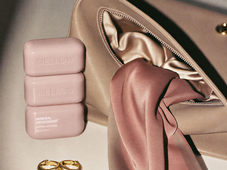 Blush pink Mellow mineral deodorant packaging styled beside a satin-lined cosmetic bag, pink fabric and gold hoop earrings.