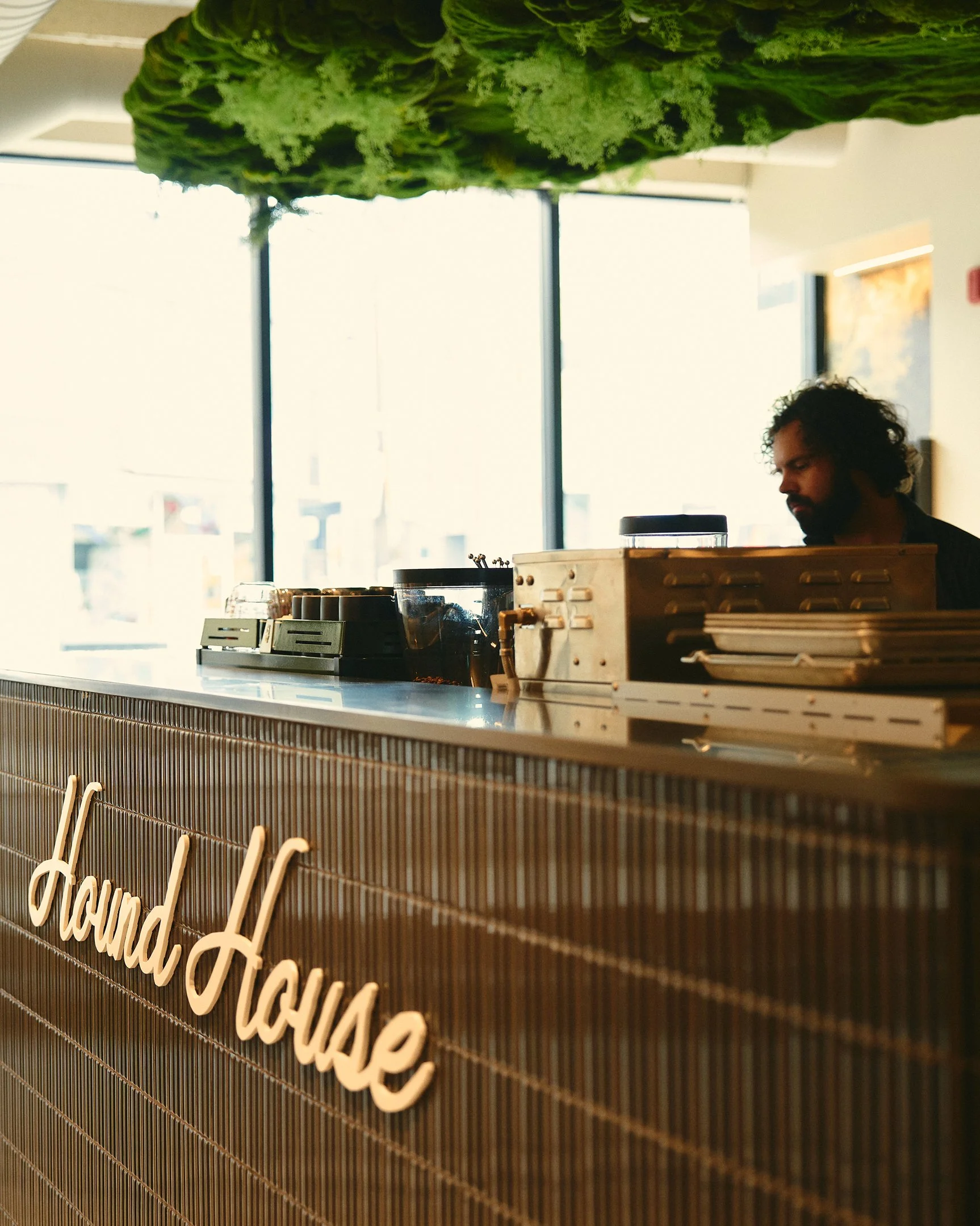 Hound House café interior with tiled service counter, warm lighting, espresso equipment, and a script-style Hound House logo, topped with lush green ceiling installation.