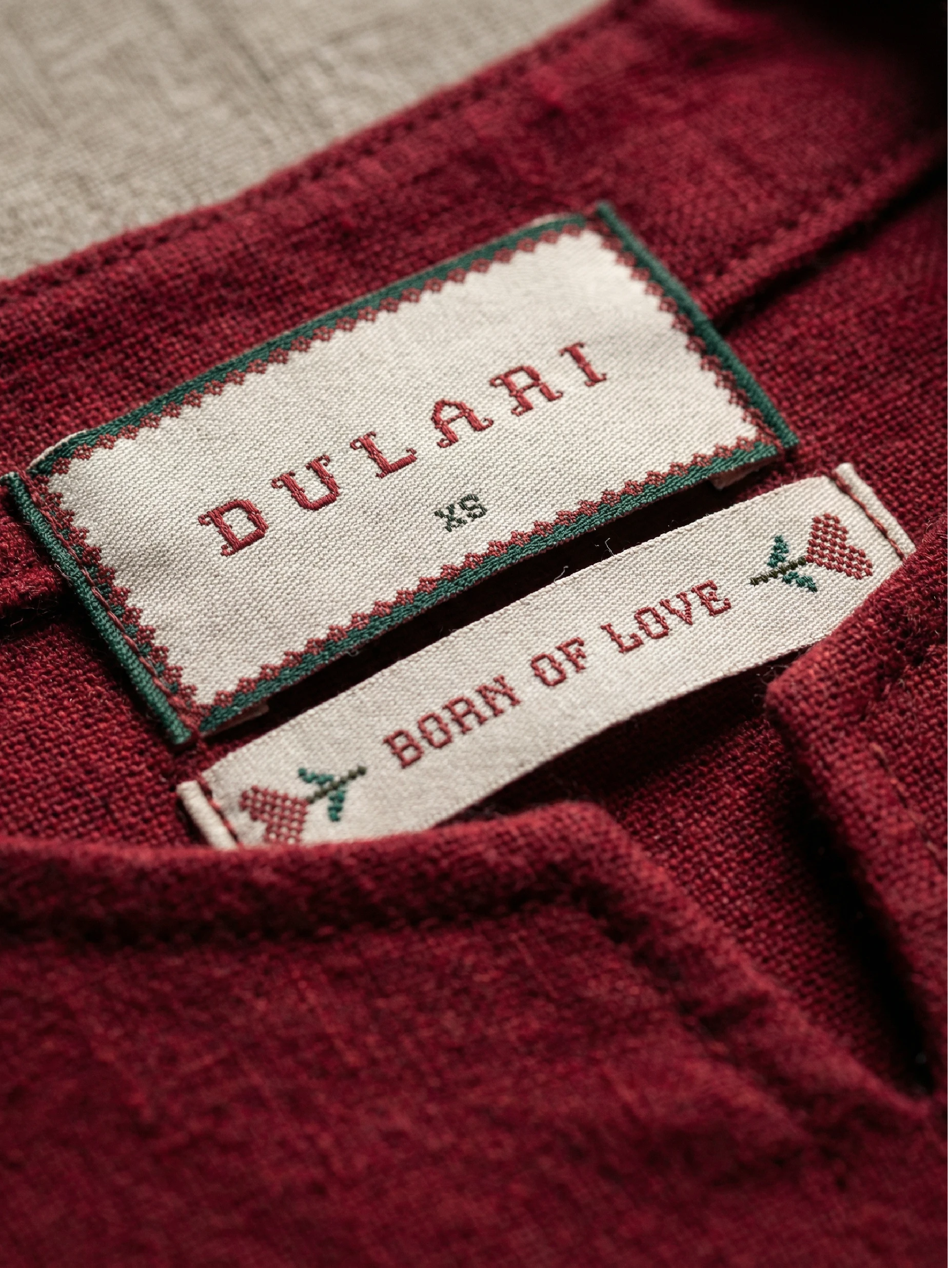 Dulari fashion brand identity woven garment labels featuring pixel-style embroidery typography, ladybug motif, and “Born of Love” tagline stitched onto textured red fabric.