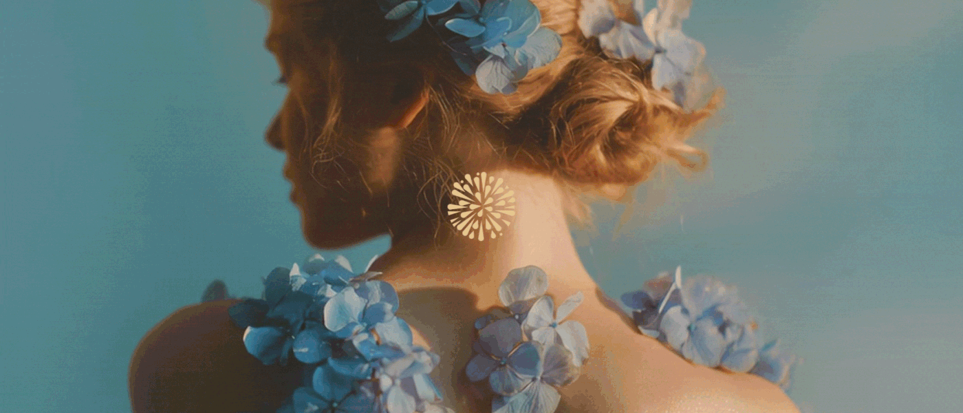 Atmospheric back-view portrait of a woman with light brown hair in a bun. Blue hydrangeas are woven into her hair and draped across her shoulders. A small, central, embossed gold mandala logo is positioned on her neck, representing the Lumere brand.