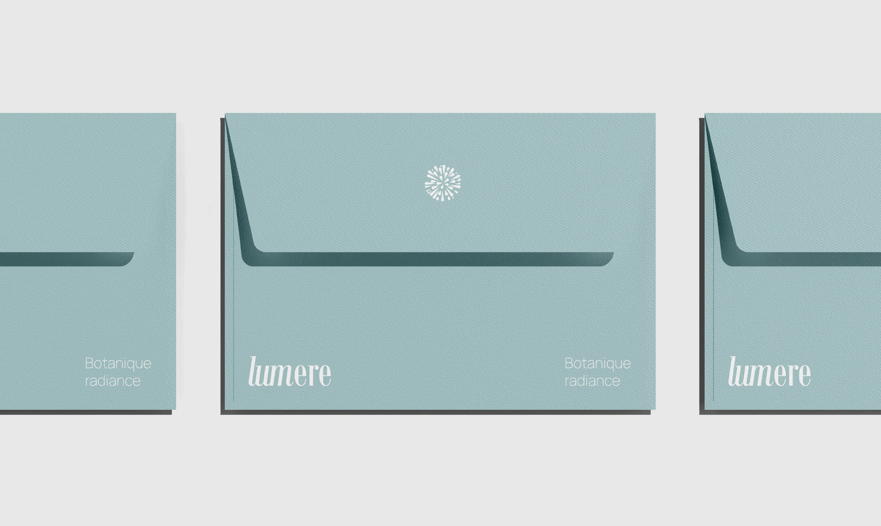 Three horizontal light-teal envelopes with a subtle paper texture. The central envelope displays a small white-printed mandala on the flap and 'lumere' and 'Botanique radiance' in a sharp serif font at the bottom corners.