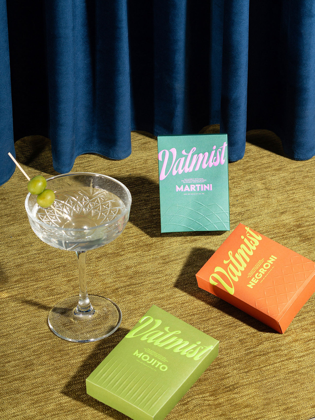 Valmist Martini, Negroni, and Mojito RTD packages arranged on a textured gold surface beside a crystal martini glass garnished with olives.