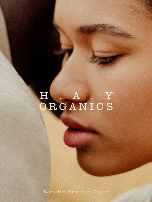 Close-up lifestyle image for HAY Organics with a soft facial profile, warm film grain, and white logo typography reading Remedies Rooted in Nature.