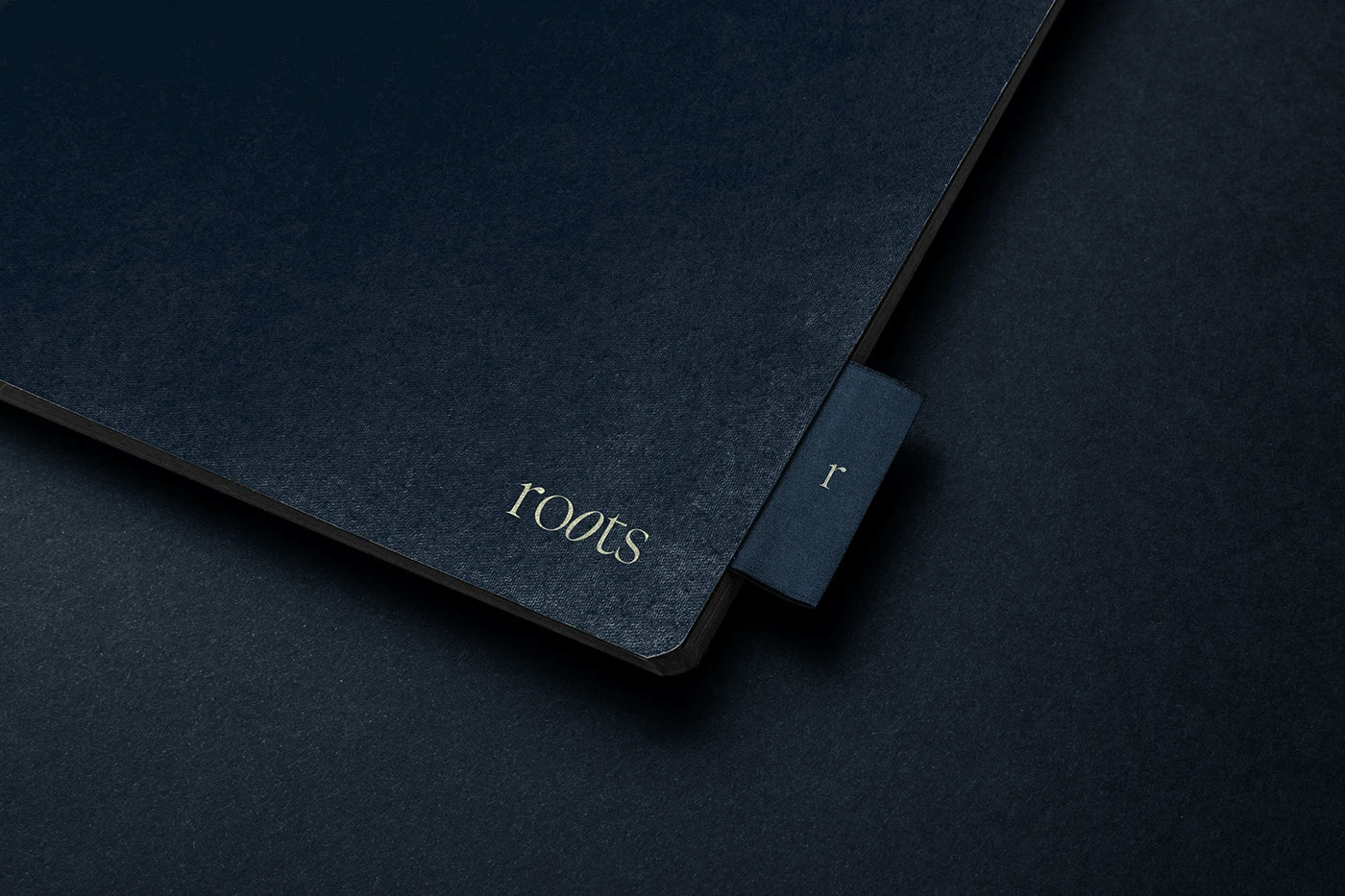 Close-up of a luxury textured navy blue notebook with the Roots logo embossed in gold foil on the cover and a small fabric 'r' tab peeking from the side.