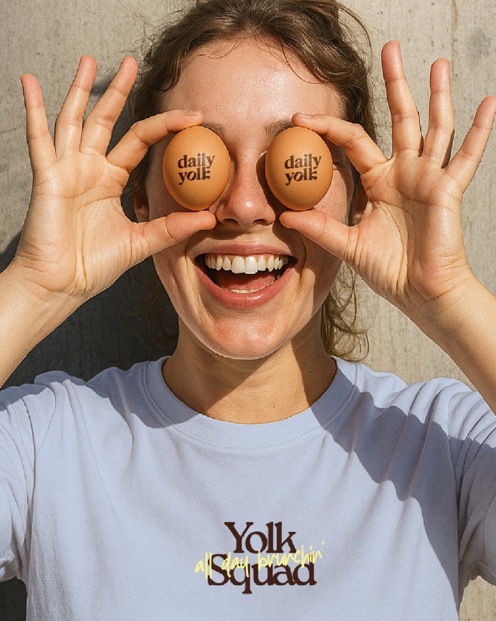 Daily Yolk lifestyle branding image showing a person holding logo-stamped eggs over their eyes while wearing a branded T-shirt with the café’s typography.