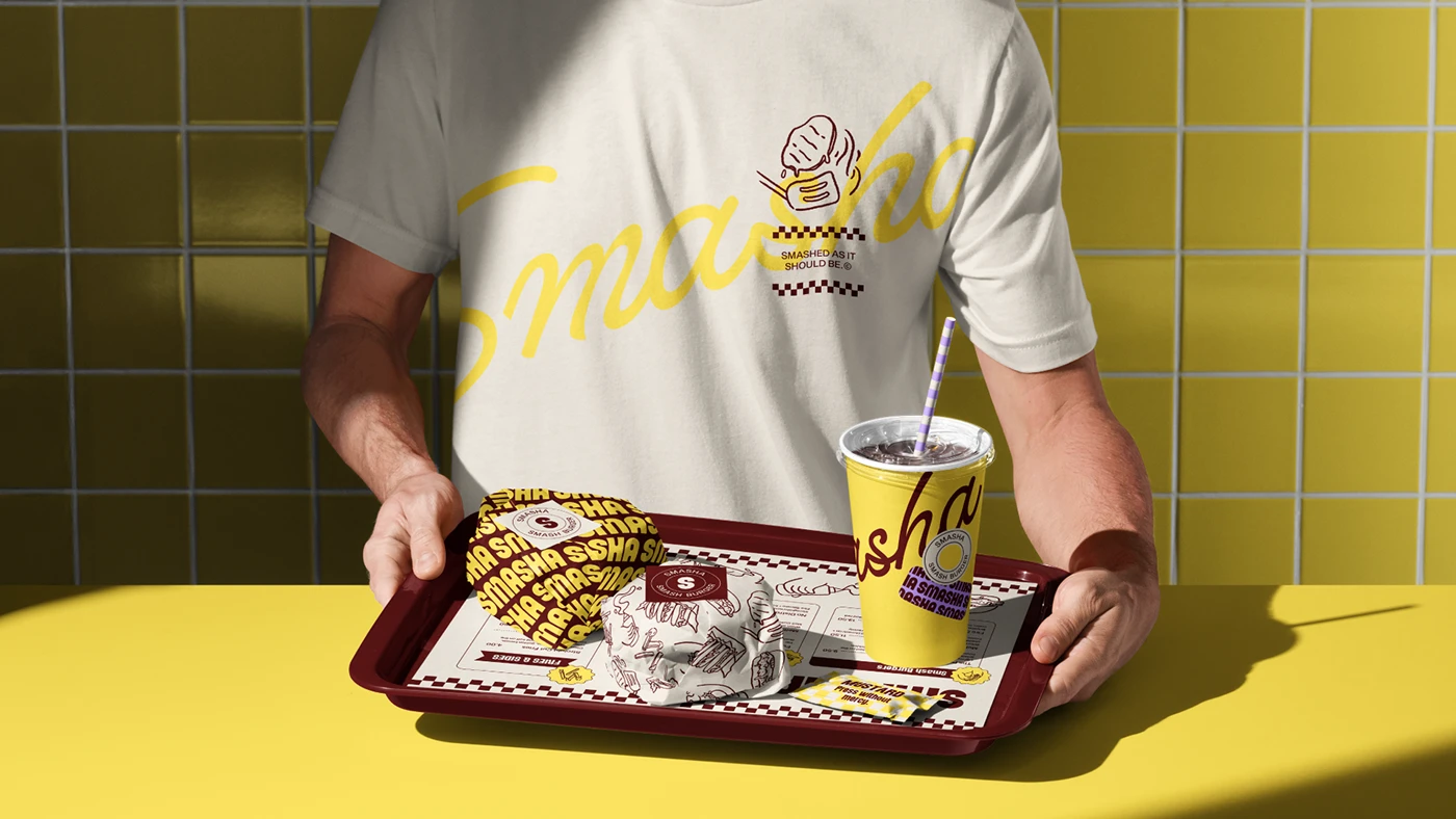 Branded Smasha staff uniform featuring a cream t-shirt with yellow script logo, paired with a tray holding wrapped burgers, branded paper liner, and a yellow drink cup against tiled walls.