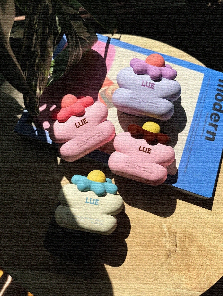 Sunlit lifestyle product scene with LUE collagen cosmetic vessels in pink, cream, and lavender arranged on a table beside graphic brand materials.
