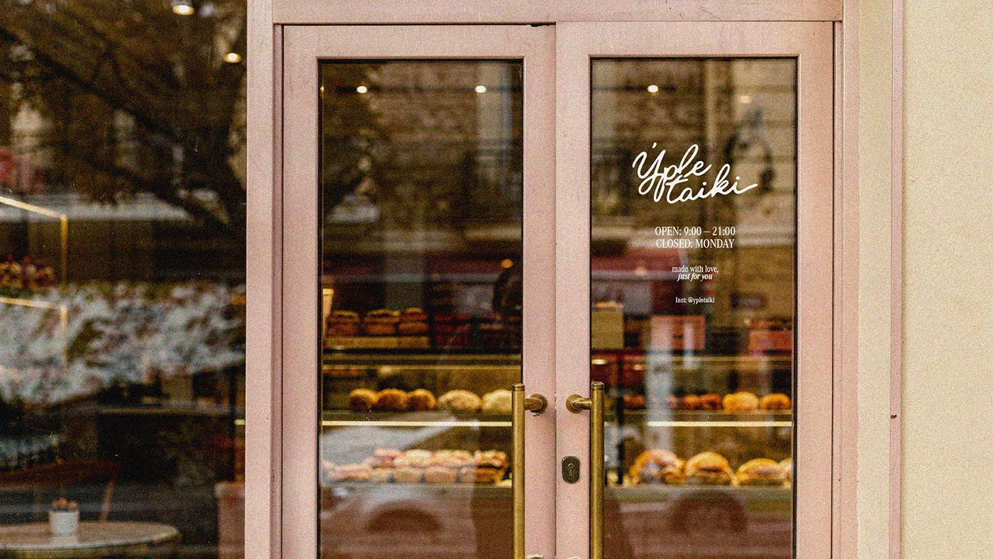 Ypletaiki café storefront with blush pink double doors and glass windows featuring script logo, opening hours, and visible pastry displays inside.