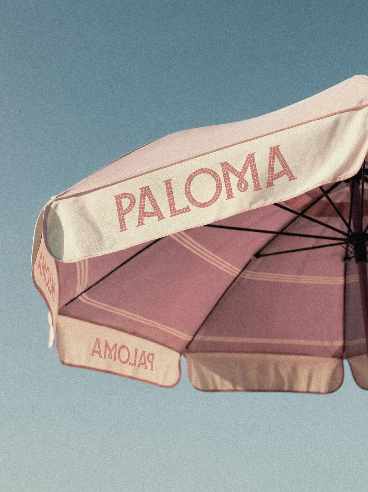 Pink and cream Paloma beach umbrella against a blue sky, featuring the mosaic-inspired resort logo as a branded hospitality application