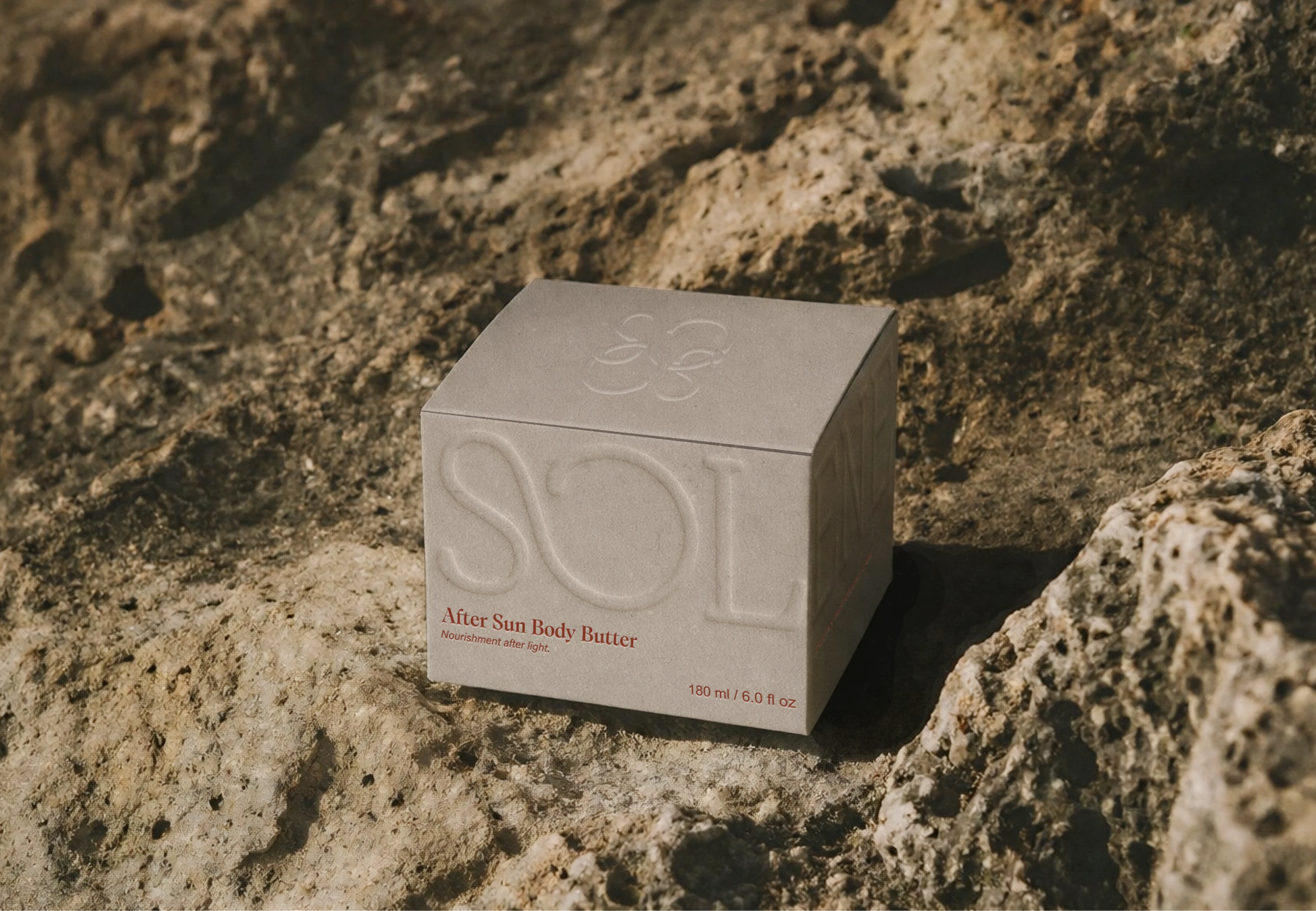 Solene SPF after sun body butter box with blind embossed logo and subtle flower mark, photographed on sunlit coastal rocks to highlight the tactile packaging design.