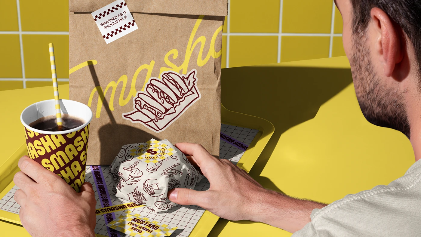 Hands reaching for a wrapped Smasha burger on a yellow tray alongside branded drink cup, mustard packet, and illustrated packaging on tiled surface.