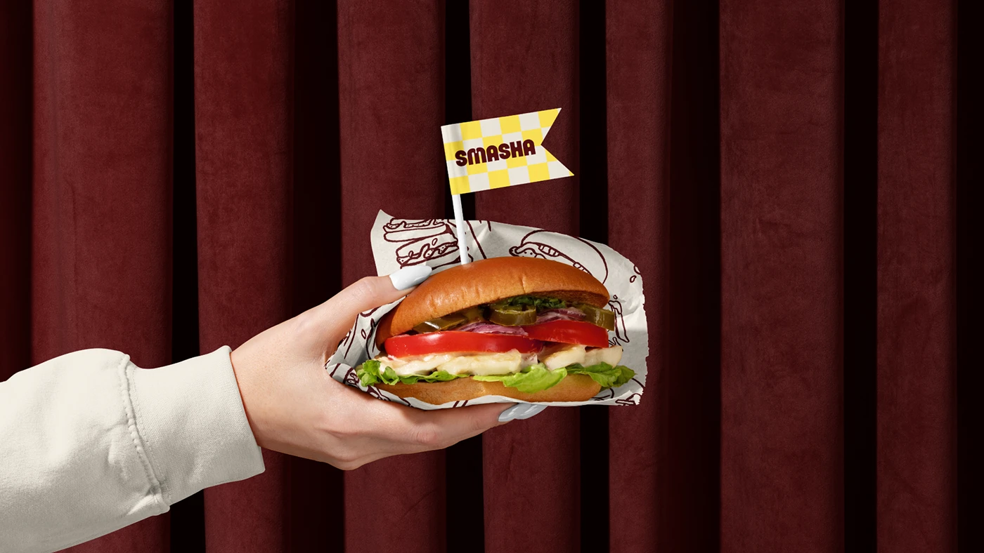 Hand holding a wrapped smash burger with a Smasha branded flag, set against a rich red background emphasizing food branding and presentation.