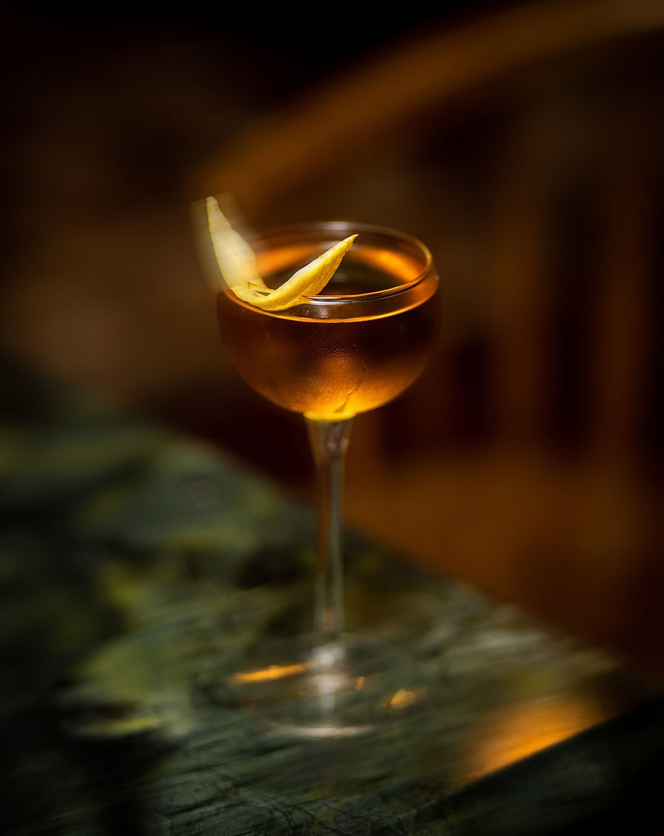 Amber cocktail with a lemon twist photographed against a warm, moody background with deep green and golden tones.