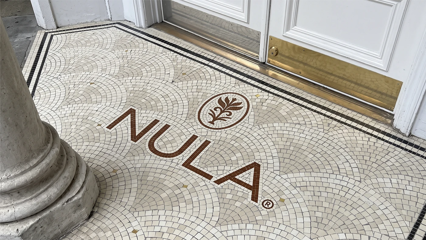 NULA brand identity applied as a custom mosaic storefront floor featuring the minimalist wordmark and botanical emblem in warm neutral tones.