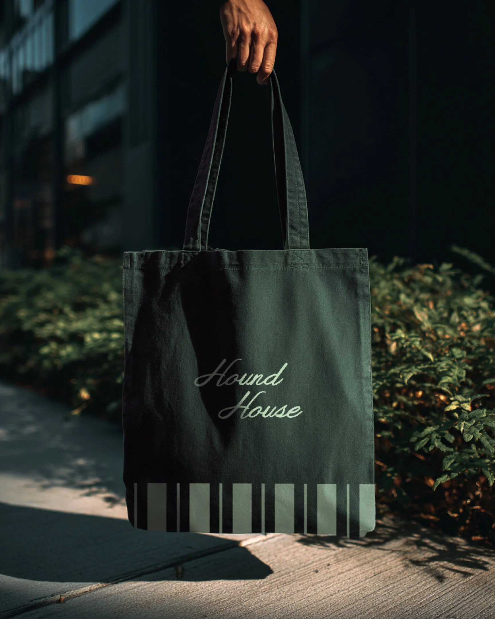 Dark green Hound House tote bag with script logo and striped detail, held by a person outdoors, highlighting branded merchandise for a modern dog café and members club.