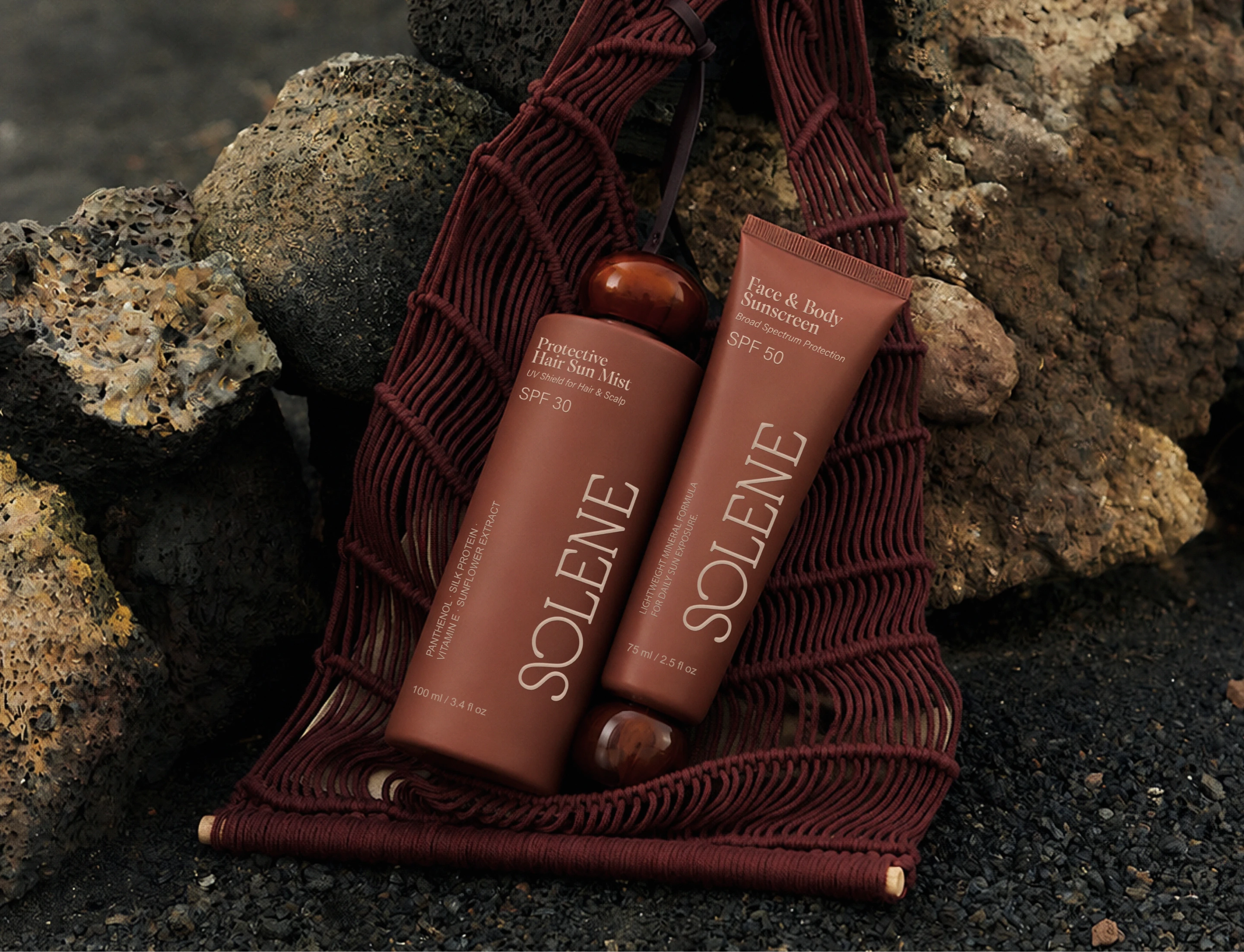 Solene SPF protective hair sun mist and face and body sunscreen displayed on a textured burgundy beach mat among rocks, showing the brand’s earthy sun care lifestyle direction.