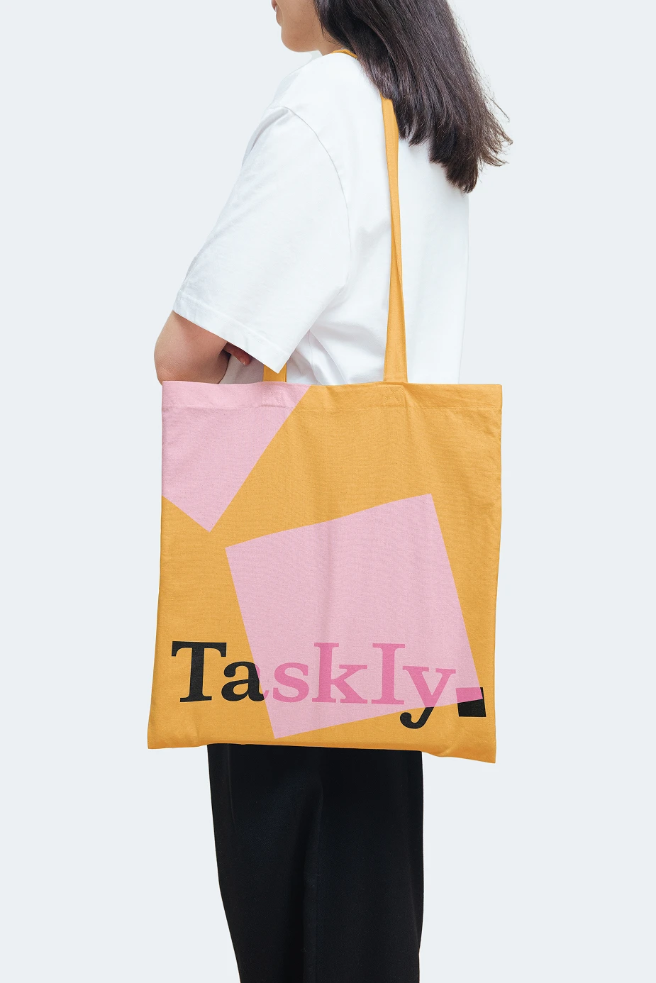 A person carrying a mustard yellow tote bag featuring the Taskly logo and oversized pink sticky-note graphics, showcasing the brand identity applied to lifestyle merchandise and modern promotional design.