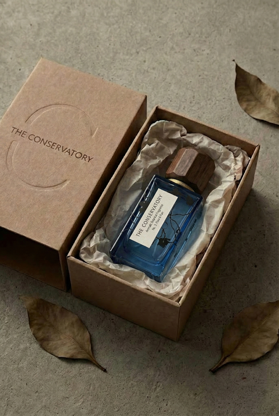 Blue glass fragrance bottle from The Conservatory presented inside a kraft paper box with tissue wrapping, highlighting premium fragrance packaging and understated brand presentation.