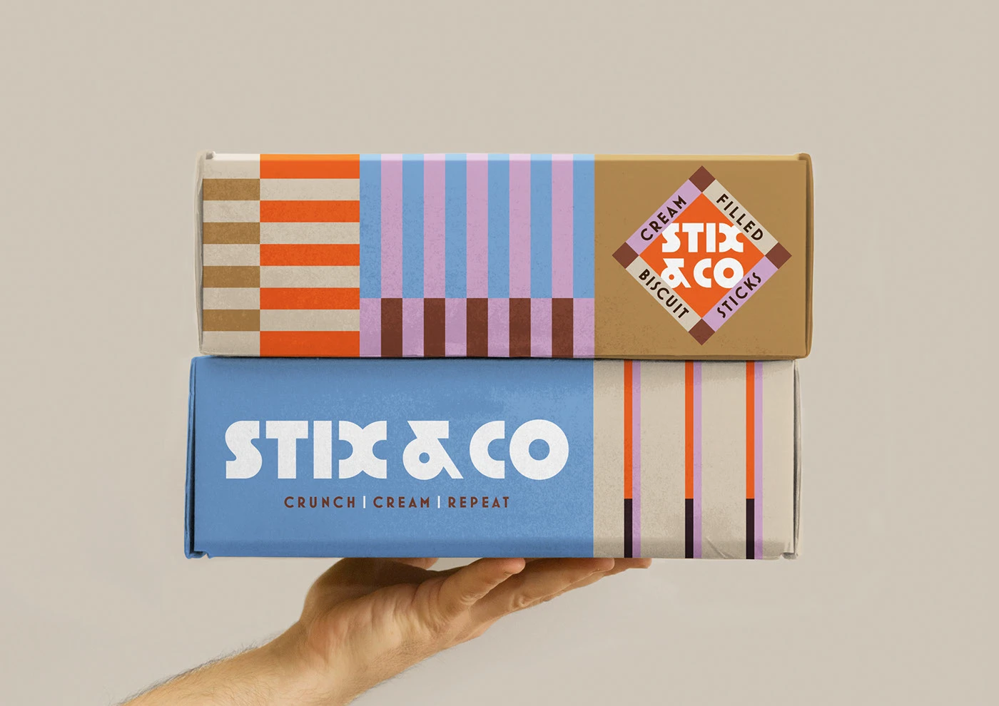 A person’s hand holding two stacked STIX & CO biscuit boxes, showcasing the side and top panel details. The top box features the diamond-shaped "Cream Filled Biscuit Sticks" seal and modular line patterns, while the bottom blue box displays the bold white brand logo and the rhythmic tagline "CRUNCH | CREAM | REPEAT."