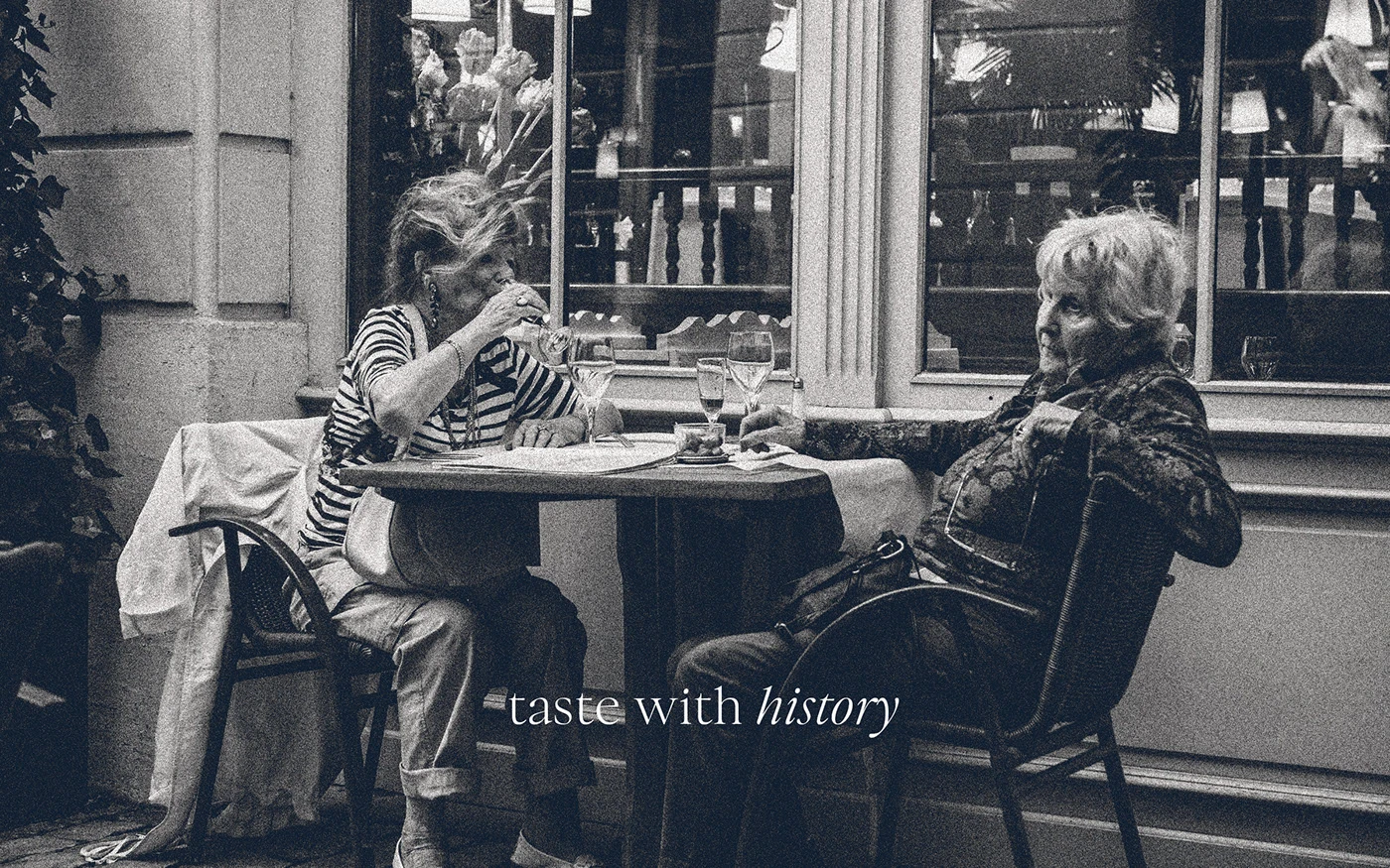 A vintage-style grainy black and white photo of two women drinking wine at a sidewalk cafe, with the brand's 'taste with history' tagline in white italicized serif.