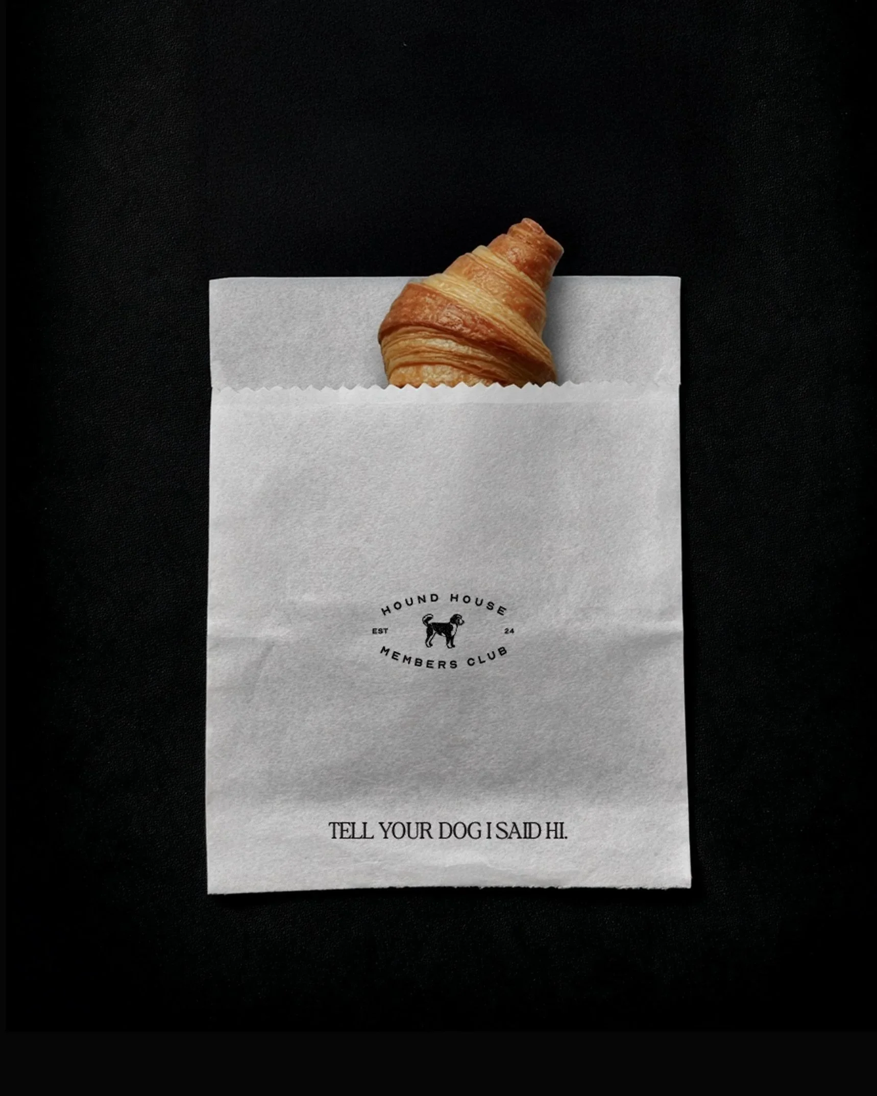 “Minimal white pastry bag branded with the Hound House Members Club logo and the phrase ‘Tell Your Dog I Said Hi,’ holding a croissant against a dark background.