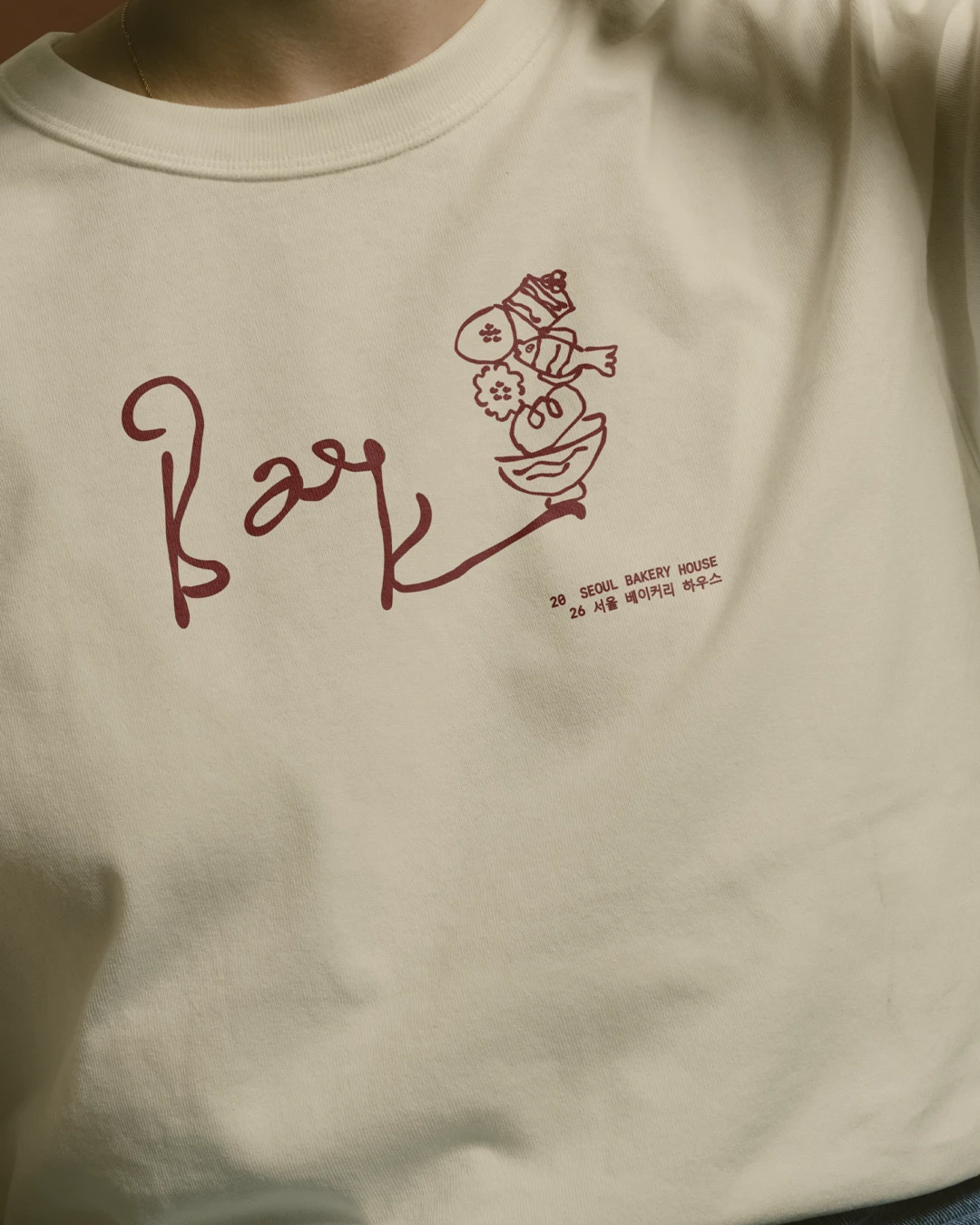 Baek Korean Bakery merchandise mockup showing a cream T-shirt printed with the deep red handwritten logo, Korean text, and playful bakery illustrations.