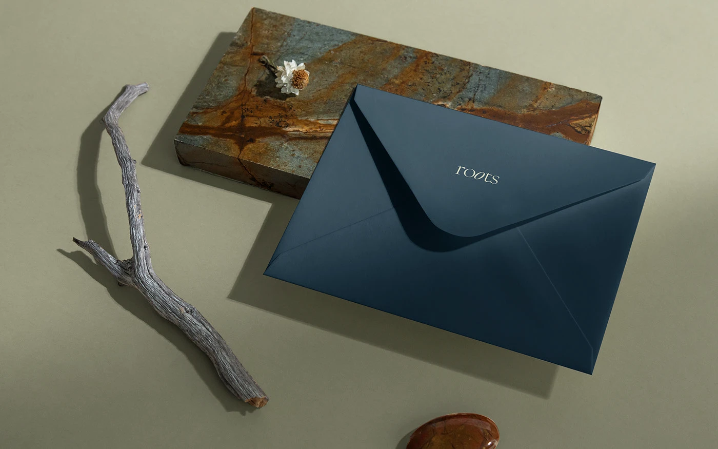 A sleek slate blue envelope with a centered Roots logo, styled in a lifestyle flat lay on a sage green surface with a decorative stone, a dried flower, and a natural wooden branch.