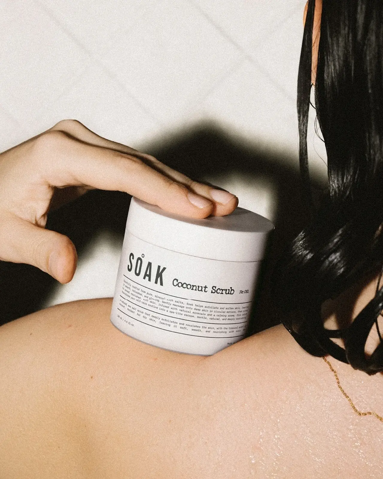 Soak Coconut Scrub jar with minimalist label design held against a shoulder in an editorial skincare lifestyle image.