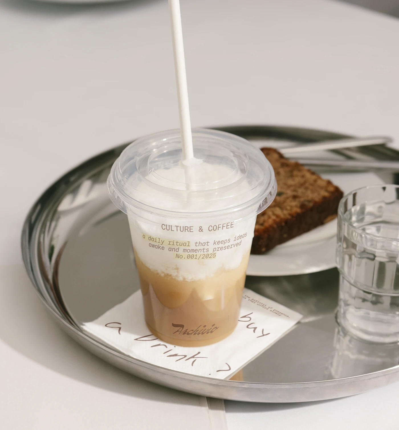 Archivio Coffee Cultura iced latte in branded takeaway cup styled on metal tray with pastry and handwritten note card reflecting archival coffee brand identity