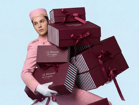 La Maison Du Lapin bakery branding campaign image featuring model in pink uniform carrying stacked burgundy pastry boxes with ribbon detailing and elegant logo design.