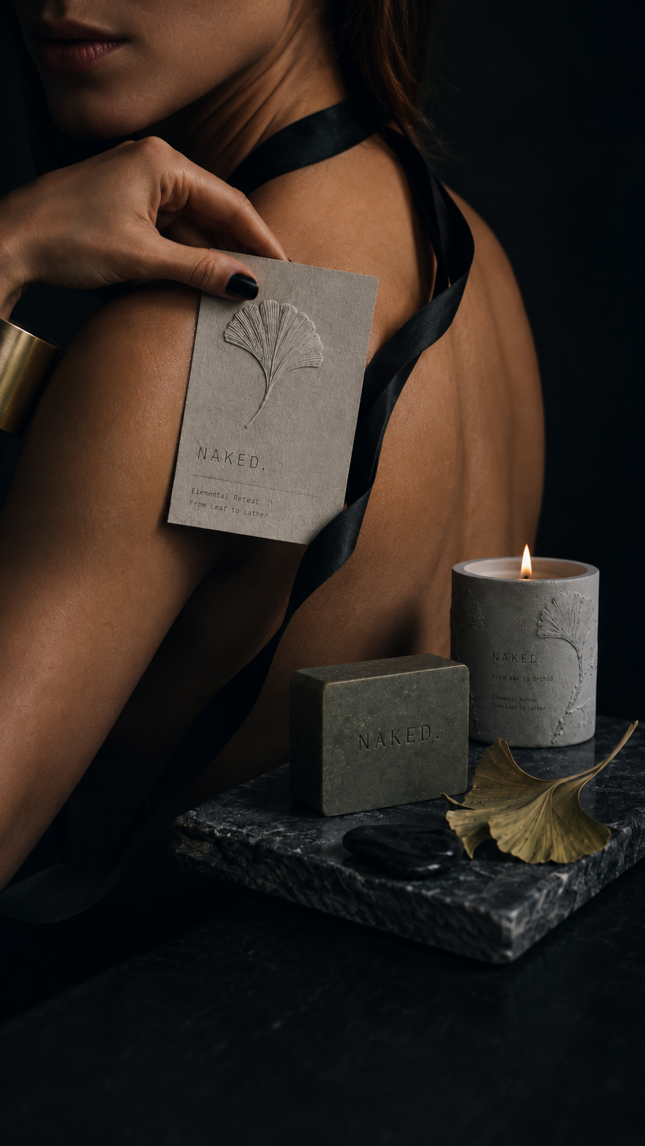 NAKED. skincare ritual scene with an embossed brand card held against skin, a green mineral soap bar, botanical candle, ginkgo leaf, and dark marble styling.