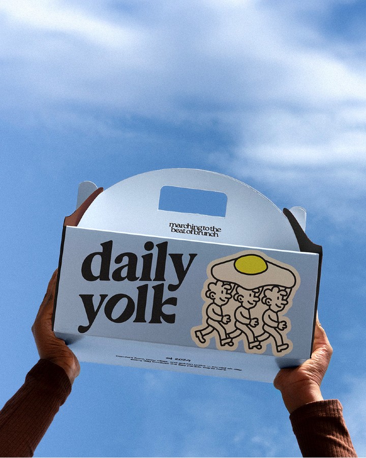 Daily Yolk brunch café packaging design showing a branded takeaway box with illustrated egg characters and bold typography held against a blue sky.