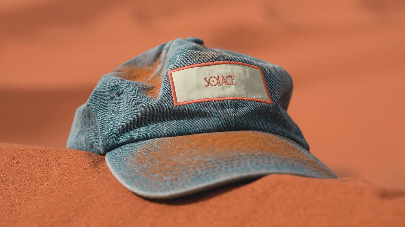 A weathered blue denim baseball cap with a 'SOLACE' embroidered patch, resting on a desert sand dune, representing the brand's connection to nature and enduring design.