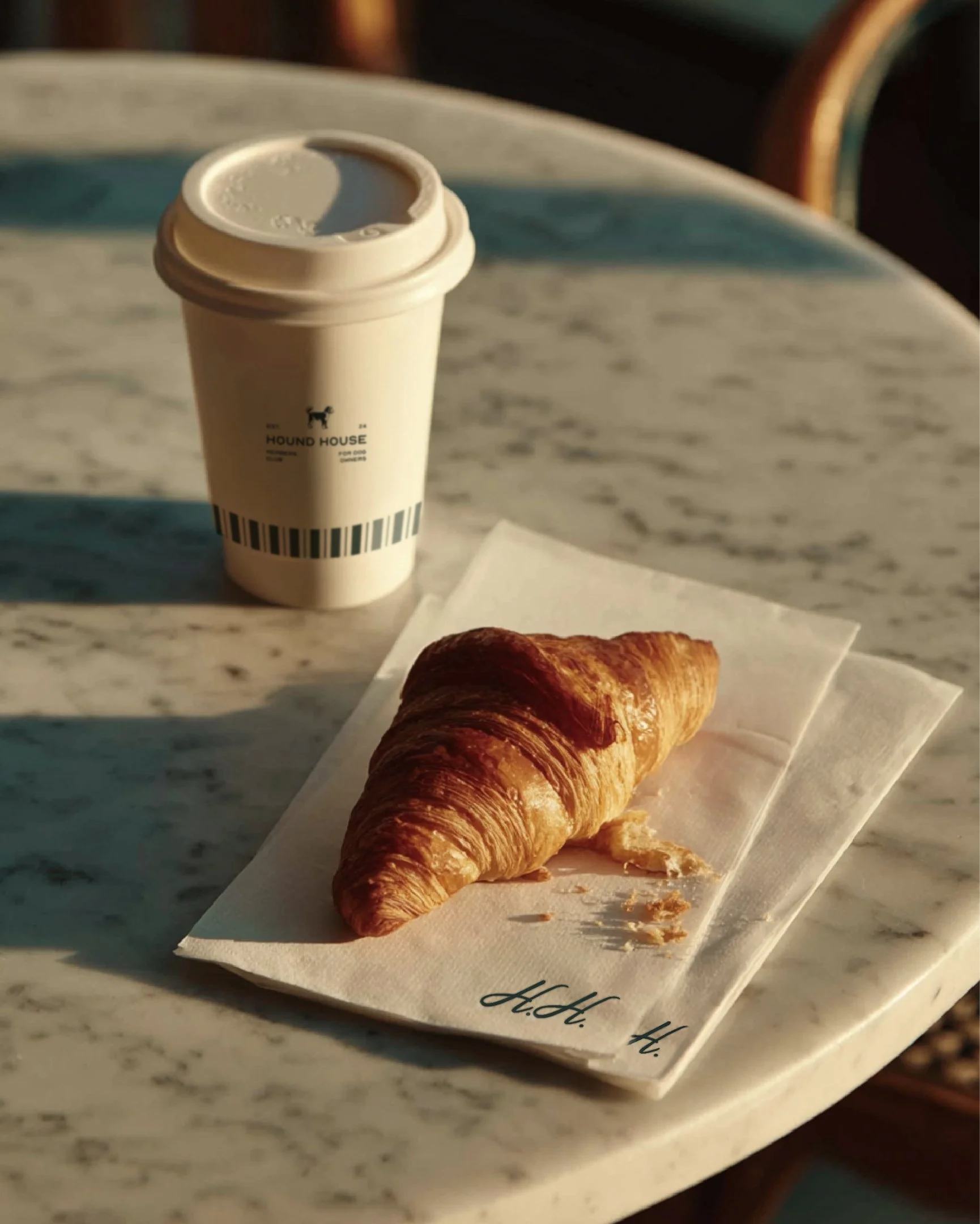 Croissant on a branded Hound House paper bag placed on a marble café table alongside a takeaway coffee cup, showcasing the dog-friendly café brand identity and minimalist packaging design.