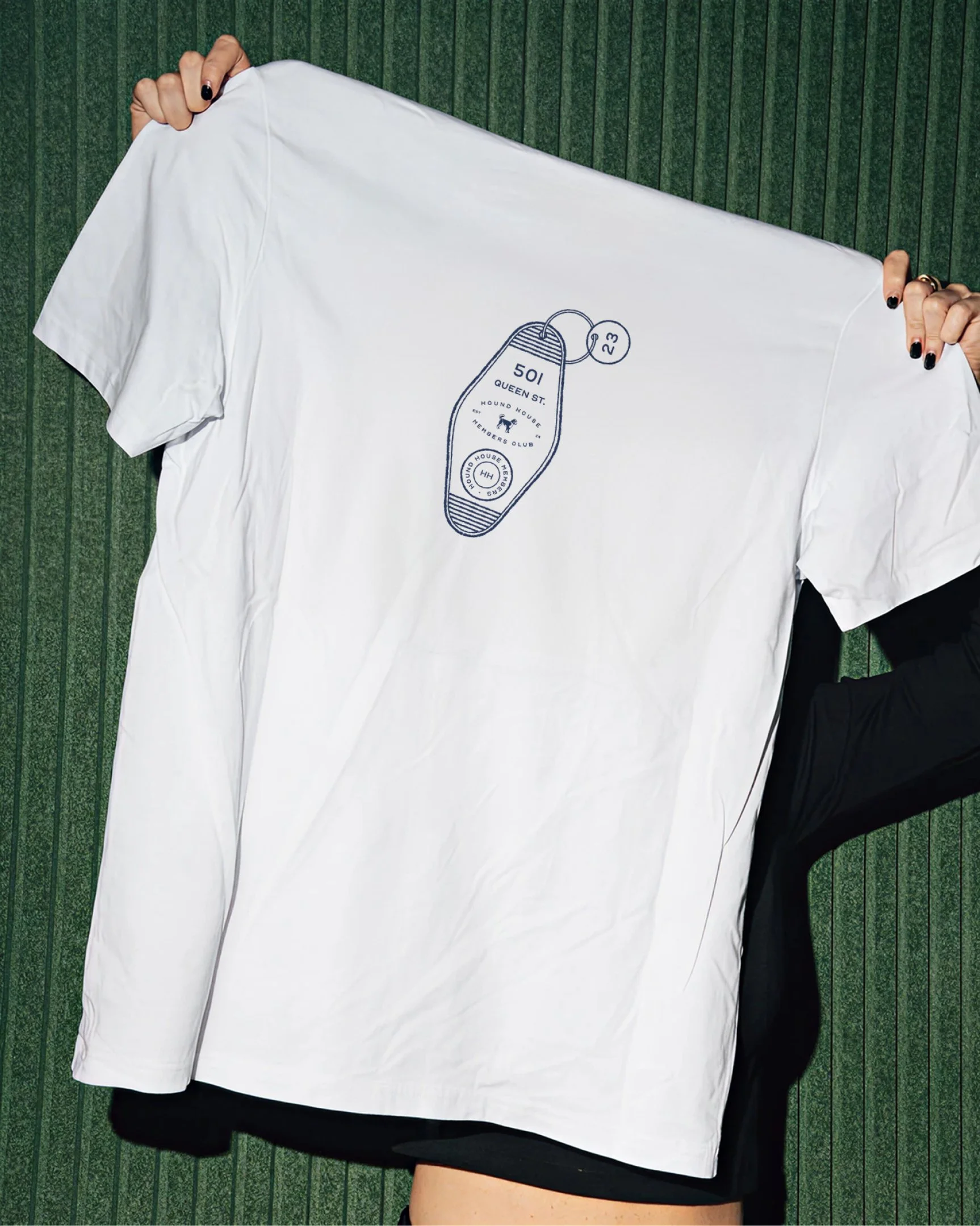 White Hound House t-shirt featuring an illustrated motel key tag graphic with address and members club branding, presented against a textured green backdrop.