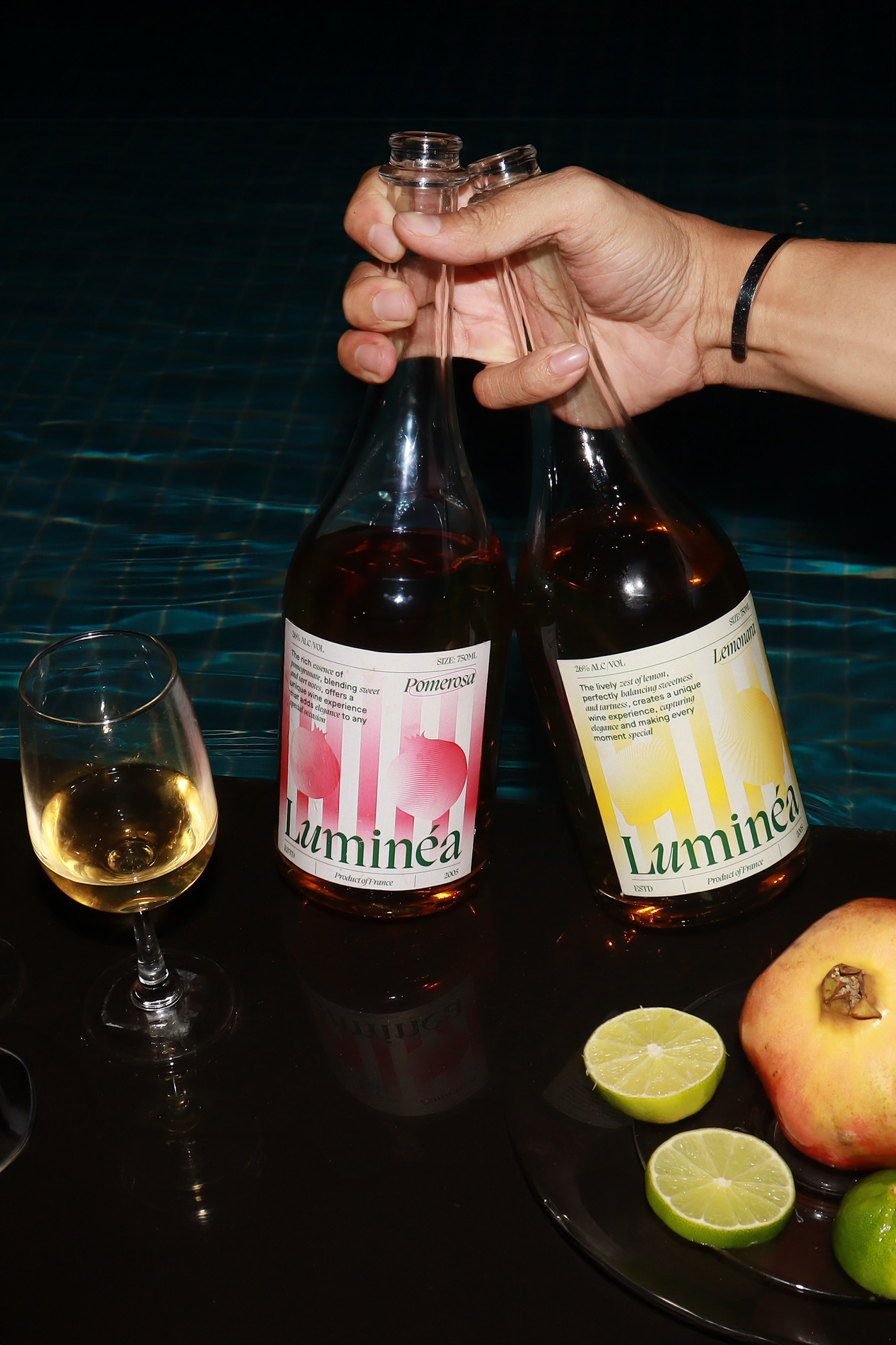 Hand holding two Luminéa Wine bottles beside a pool, displaying the gradient label designs in pink and yellow with premium editorial typography and modern beverage packaging details.