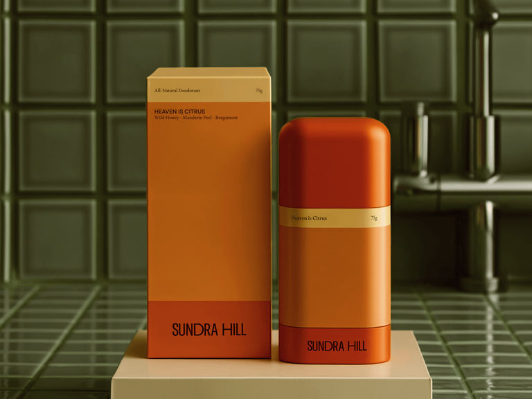 Sundra Hill deodorant packaging design by Studio One Eighty featuring a bold orange cylindrical deodorant and matching minimalist box with clean typography, photographed on a green tiled bathroom surface — a modern, architectural approach to natural body care branding.