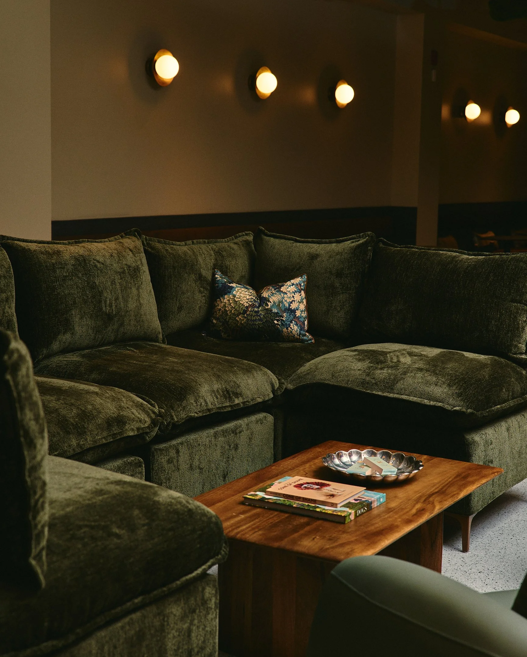 Lounge-style seating area with plush green sofas and warm lighting, illustrating the interior atmosphere of a private pet social club designed for dogs and their humans.