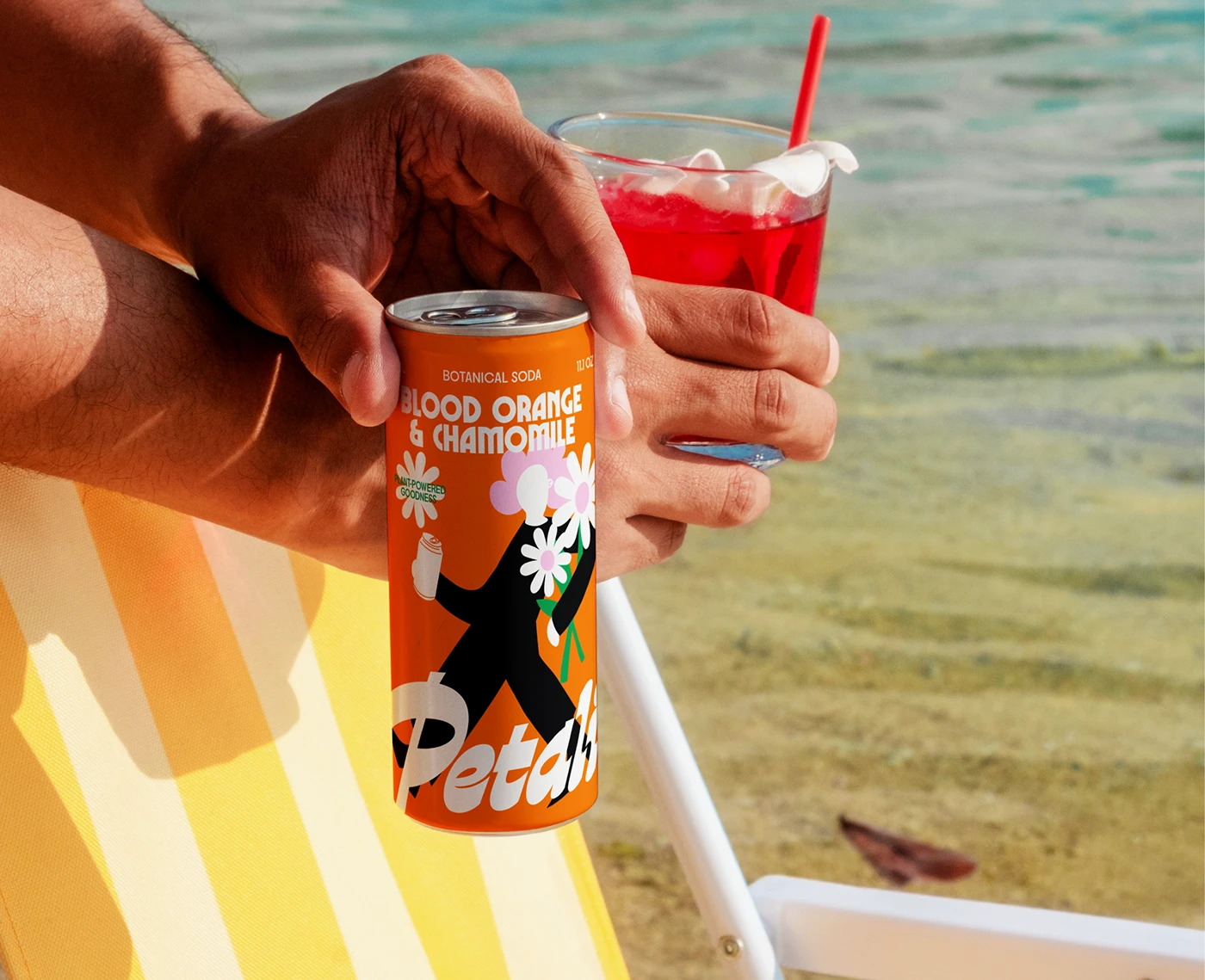Petali Soda can held beside a red drink near the beach—vibrant beverage packaging design featuring dopamine orange color and groovy 70s-inspired illustration.