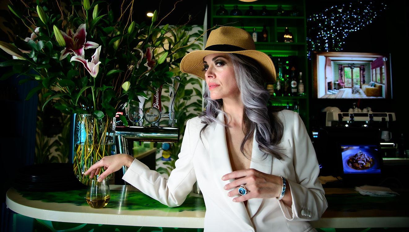 Woman with grey hair in a white blazer and tan fedora at a bar