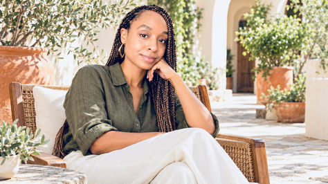Dr. Abdullah sits on a wicker chair in a sunny courtyard, wearing a green shirt and white pants, smiling softly.