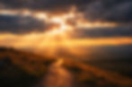 Golden sunset rays break through dark clouds over a winding hillside path and distant mountains, creating a dramatic glow.