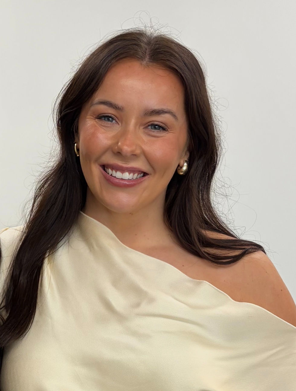 Portrait photo of a smiling woman with long dark hair, wearing a cream one-shoulder top and dark trousers, on a light background.