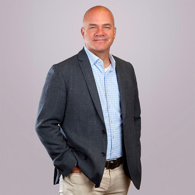 Headshot of a bald man with his hand in his pocket in a suit on a gray backdrop