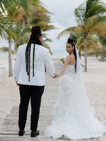 Person in tuxedo and bride walk sandy path between palm trees.