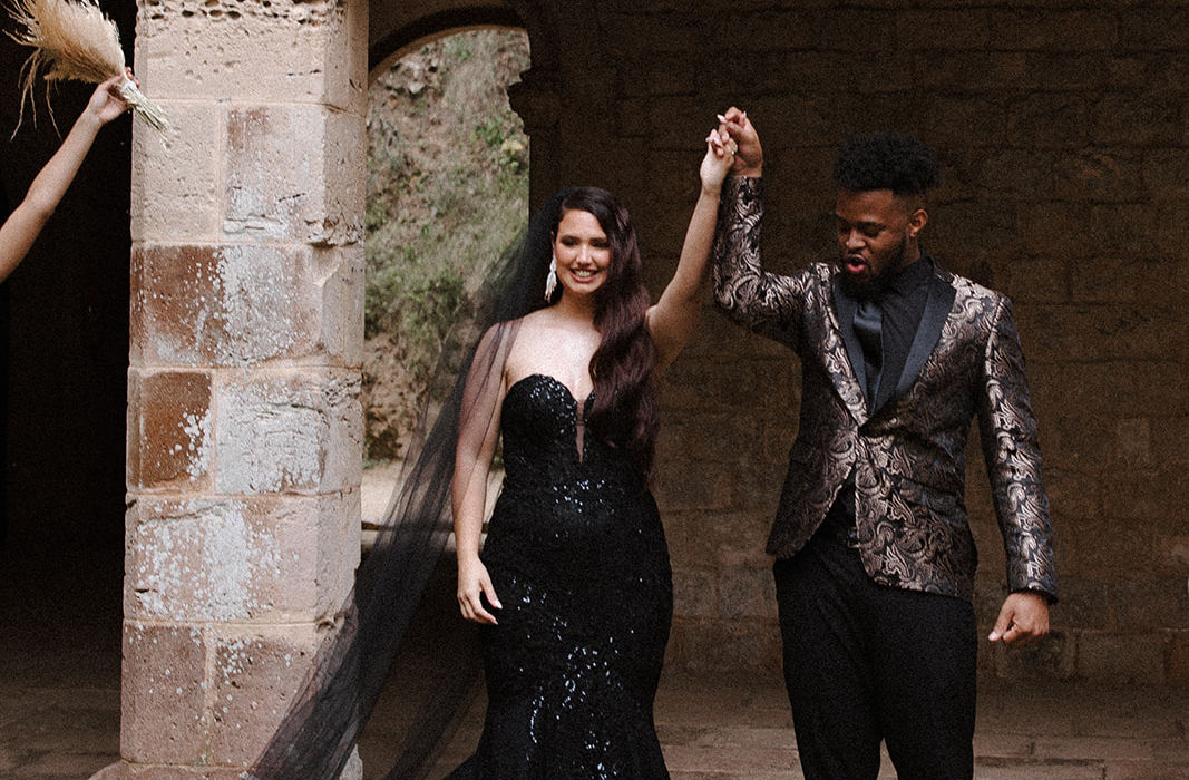 Couple in black wedding attire holding hands under stone archway celebration.