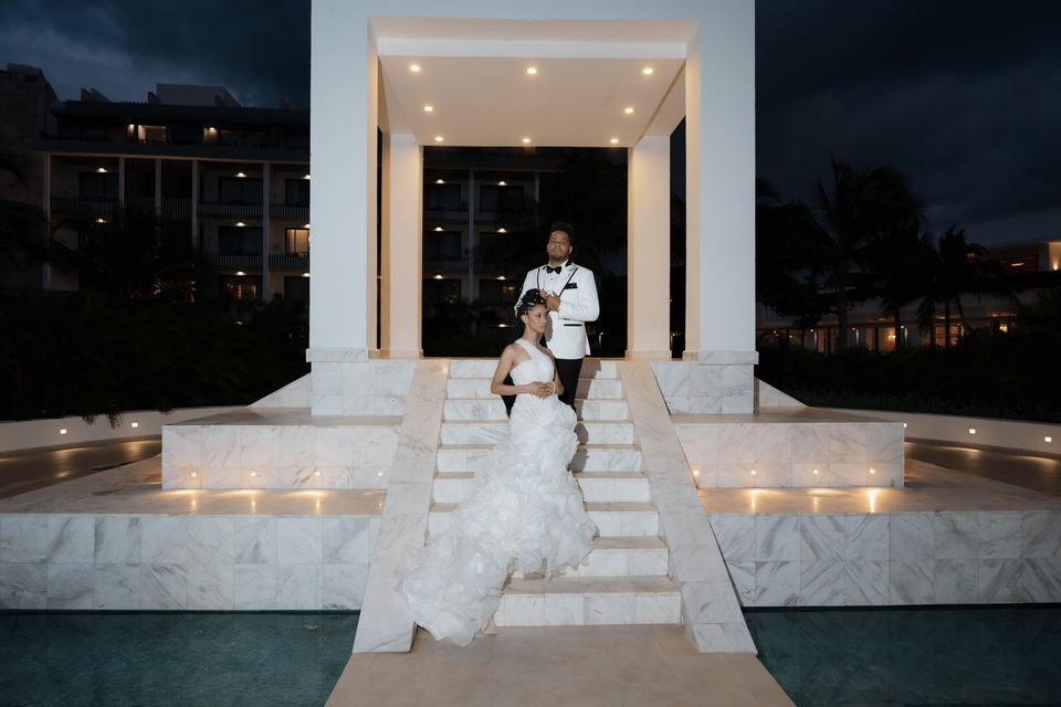 Newlyweds posing on elegant white marble steps at night, resort background.