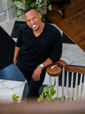 Interior designer standing on a staircase in a stylish home featuring modern black-and-white flooring, wood railing, and decorative greenery.